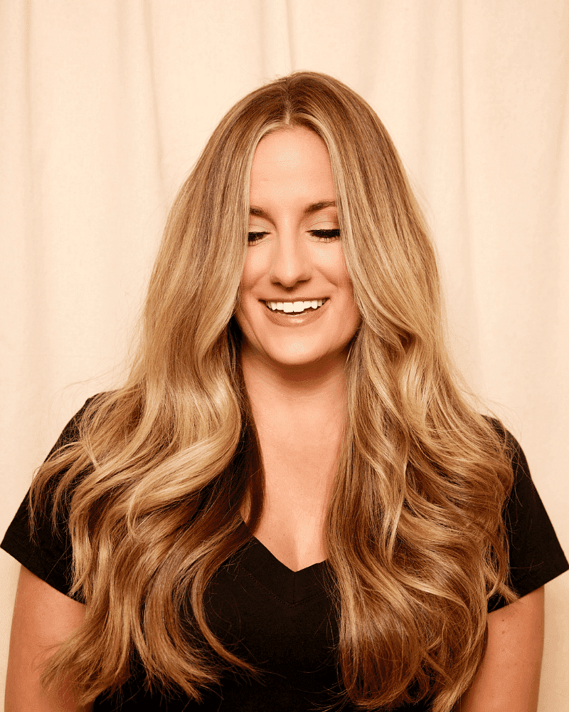 Smiling woman with long blonde wavy hair, wearing a black top, against a beige background.
