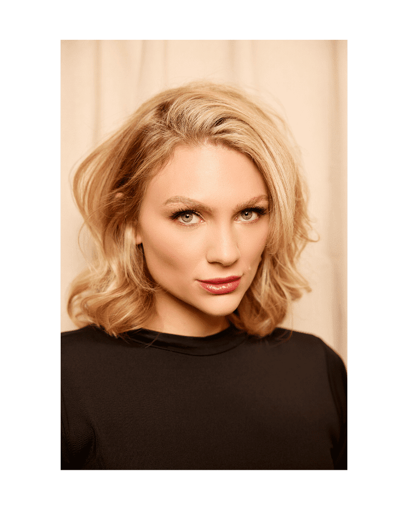 Portrait of a woman with blonde hair and a black top, posing against a beige background.