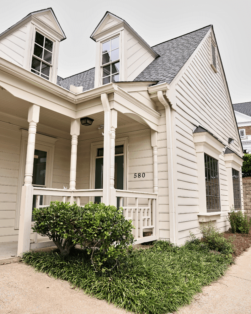 Charming beige house with a porch, twin dormer windows, house number 580, and green shrubbery.